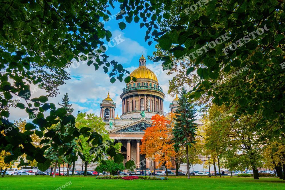 На фотографии изображён Исаакиевский собор в Санкт-Петербурге, видимый сквозь густую листву осенних деревьев. Золотой купол собора сверкает на фоне голубого неба, окружённый осенней палитрой красок. Зелёные и жёлтые листья создают естественную рамку вокруг величественного здания, подчёркивая красоту осеннего Петербурга.